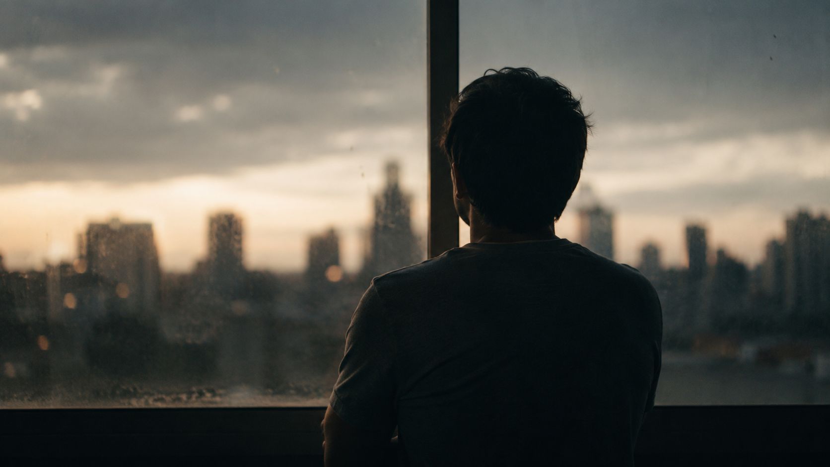 A silhouette of a man looking out of a large window at a sunset city skyline.